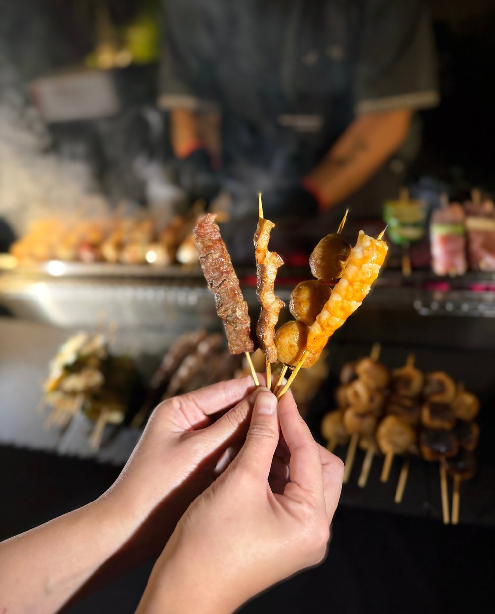 Hand holding beef, pork, meatball and shrimp yakitori skewers in front of the grill